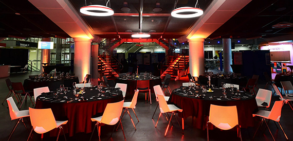 Atrium set for a dinner with round tables, orange chairs and red uplighting beside a staircase.