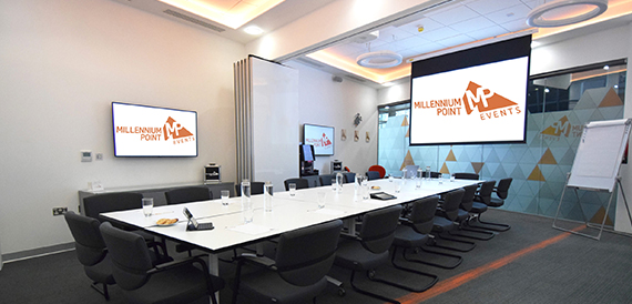 Boardroom with a long white table, black chairs and two wall-mounted screens.