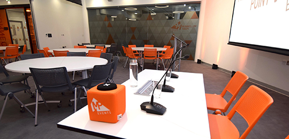 Breakout area with round tables, orange and black chairs and a projector screen.