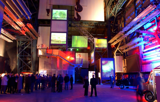 Industrial exhibition hall lit in red and blue with visitors gathered beneath suspended screens