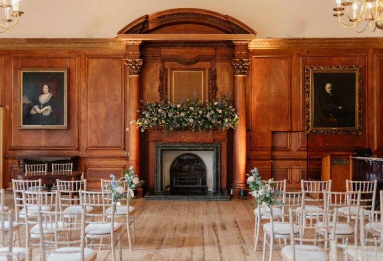 Wood-panelled room set for a ceremony with gold chairs and a floral fireplace display.