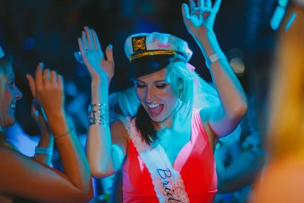 Bride-to-be in a captain's hat dancing with friends under blue lighting
