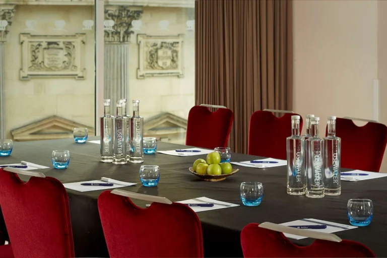 Boardroom table set with water bottles and green apples, flanked by red velvet chairs.