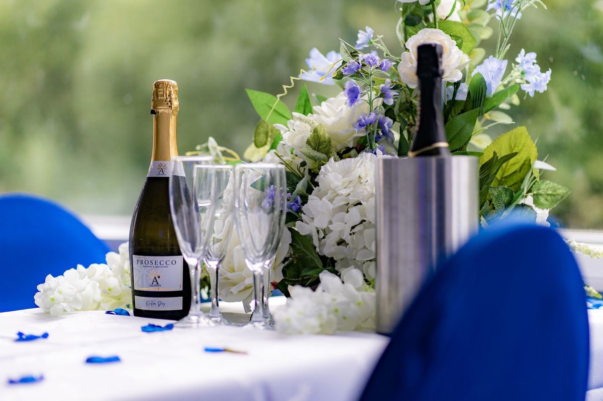 Prosecco and glasses set beside white and blue flowers with blue chair covers