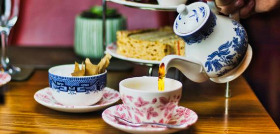 Afternoon tea being poured from a patterned teapot into floral cups on a wooden table