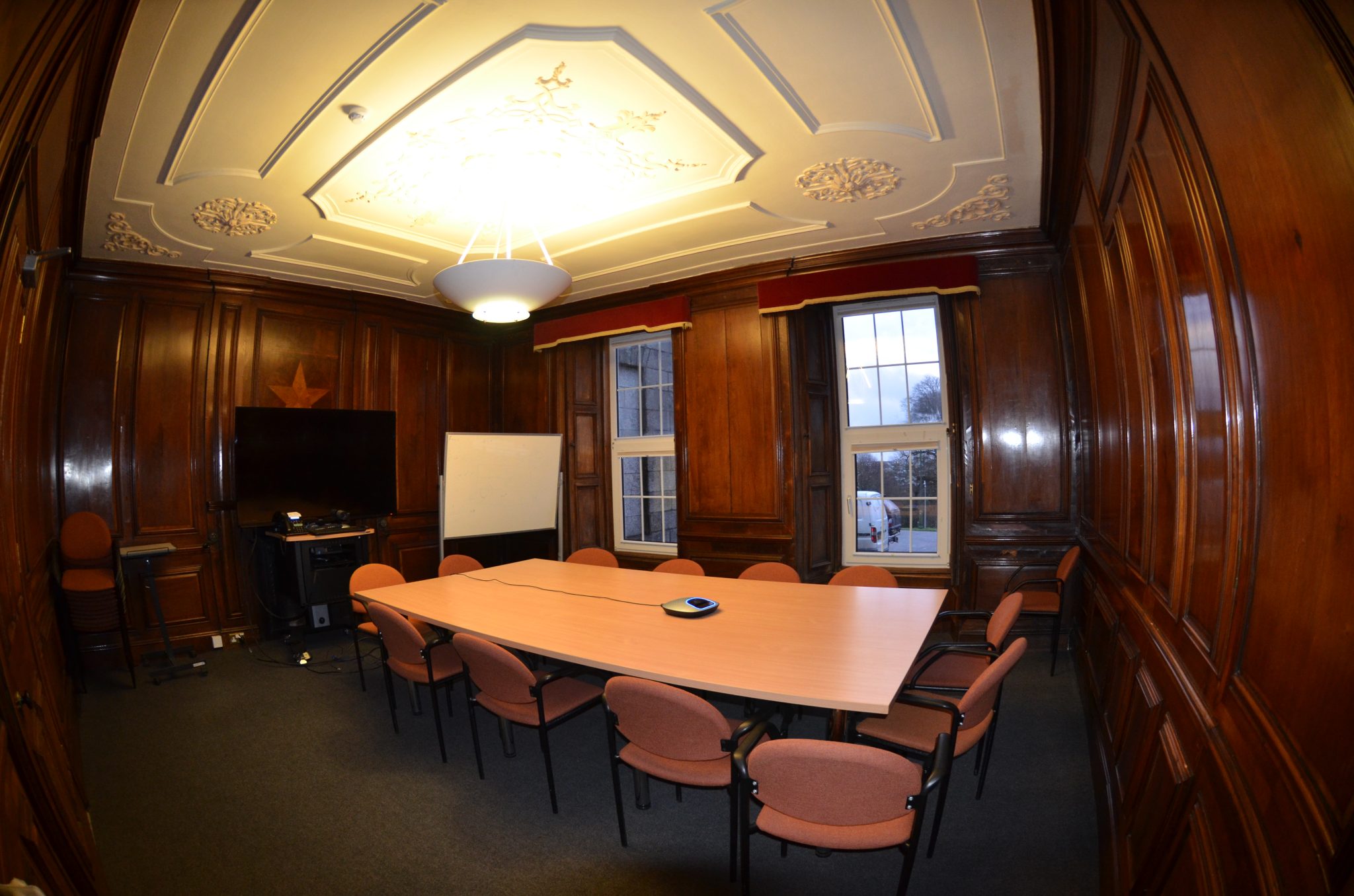 Wood-panelled boardroom with a long table, orange chairs and a wall-mounted screen.