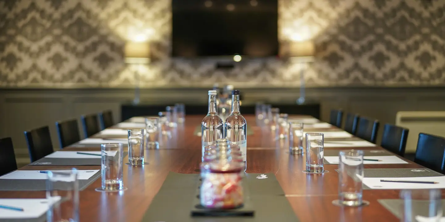 Boardroom table set with water bottles, glasses and notepads facing a wall-mounted screen
