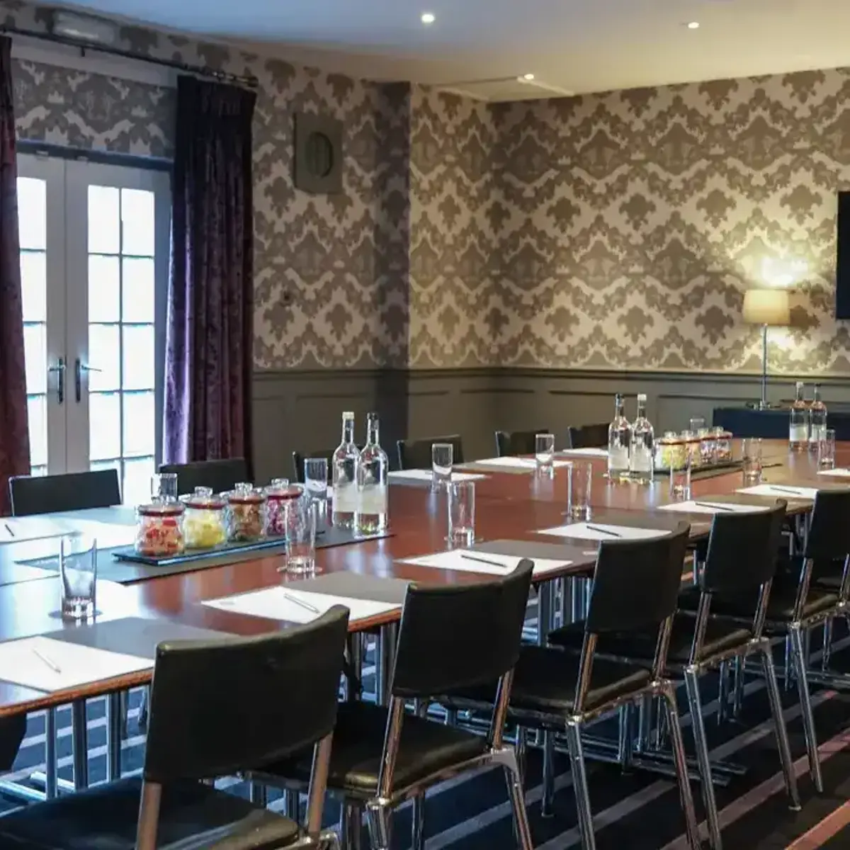 Boardroom table with water bottles, sweet jars and glasses beneath patterned wallpaper