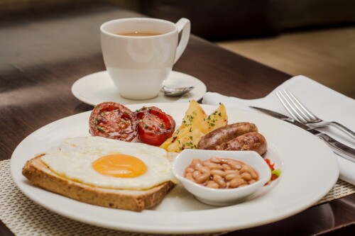 Breakfast plate with fried egg, tomatoes, beans and sausages beside a cup of tea.